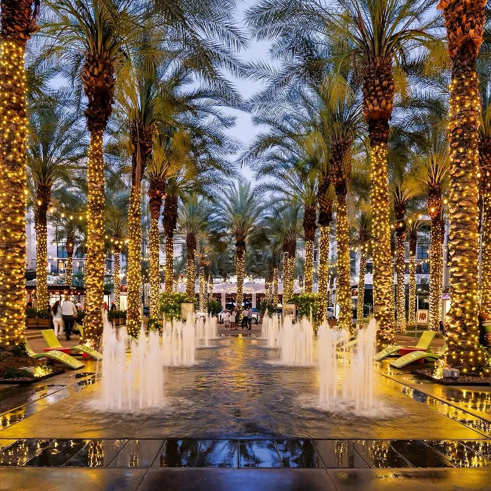 A path lined with tall palm trees wrapped in glowing string lights leads to a vibrant plaza with fountains spurting water. People walk and lounge in chairs, surrounded by the illuminated ambiance. Twilight sky above enhances the scene.