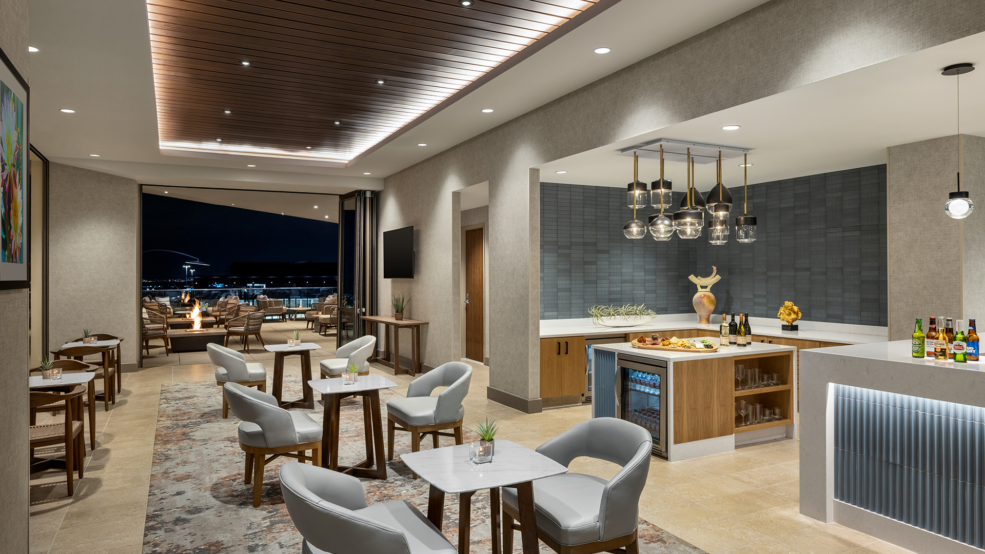 A modern lounge with gray chairs and wooden tables, featuring a bar area with hanging lights and a lit display of bottles. The room opens to a balcony with a view of the cityscape at night. The space has a warm, inviting atmosphere.