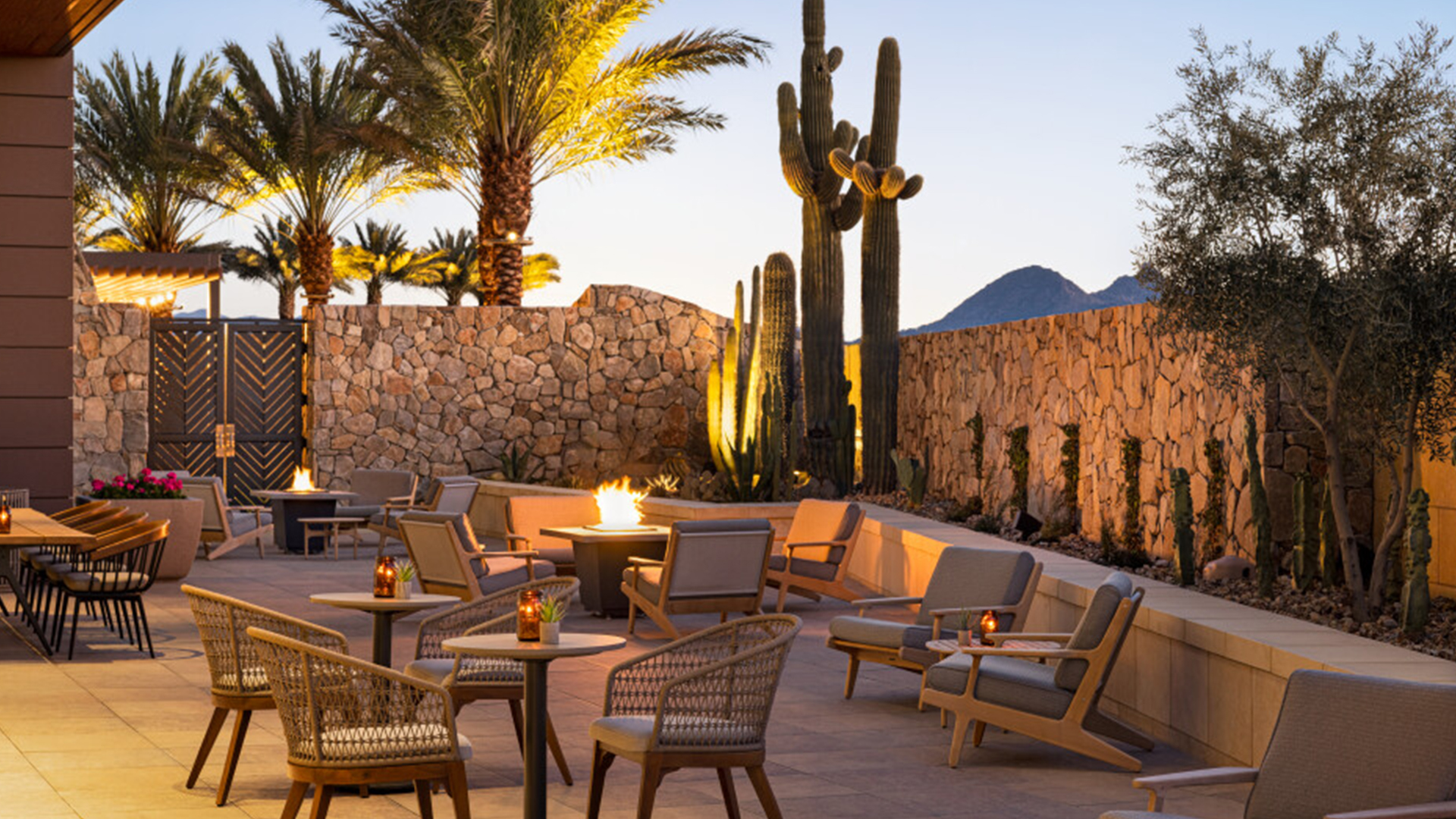 A cozy outdoor patio at sunset featuring wicker chairs and tables surrounding warm fire pits. Tall cactus plants and palm trees are in the background, next to a stone wall, with soft lighting creating a relaxing atmosphere.