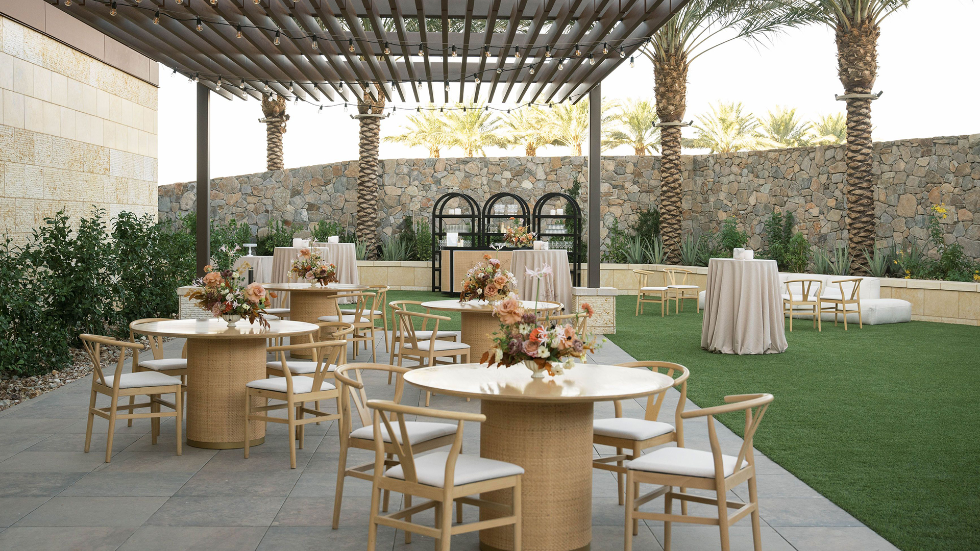 An outdoor event space with wooden tables and chairs arranged on tiled flooring and artificial grass. Each table is decorated with floral centerpieces. A stone wall and palm trees are in the background, with cocktail tables scattered around.