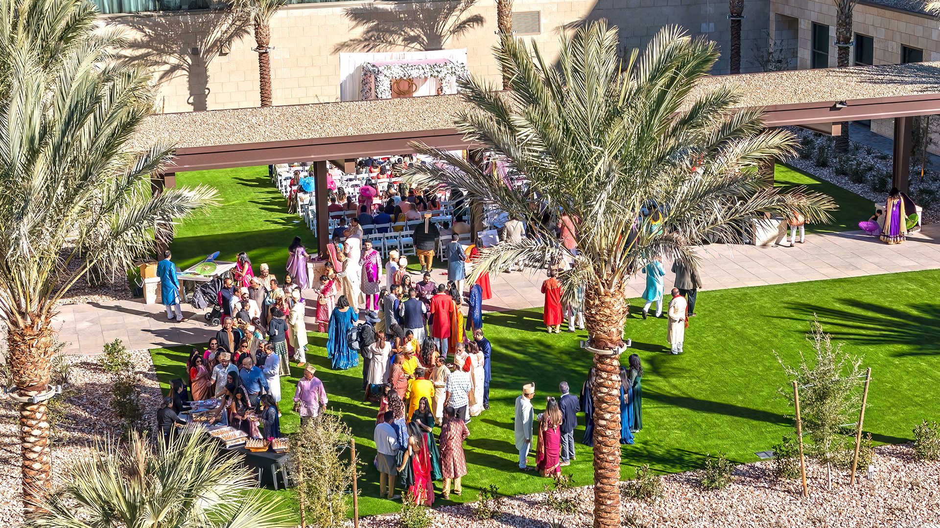 Aerial view of an outdoor event on a green lawn with people in colorful traditional clothing. Palm trees border the gathering, and tables are arranged under a gazebo. A building is visible in the background.