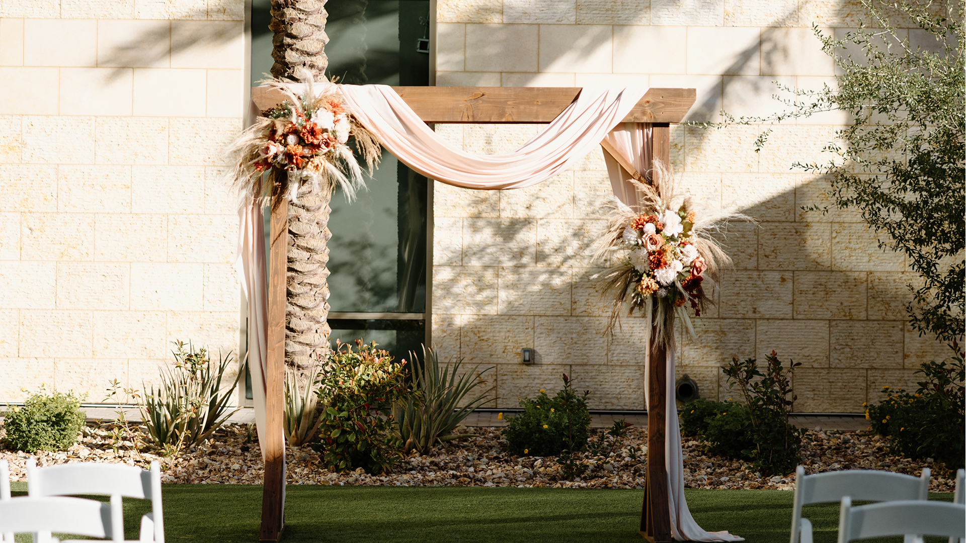 A wooden wedding arch draped with soft beige fabric and adorned with floral arrangements, featuring white and orange flowers and pampas grass, stands outdoors on a green lawn. The background shows a stone wall and plants.