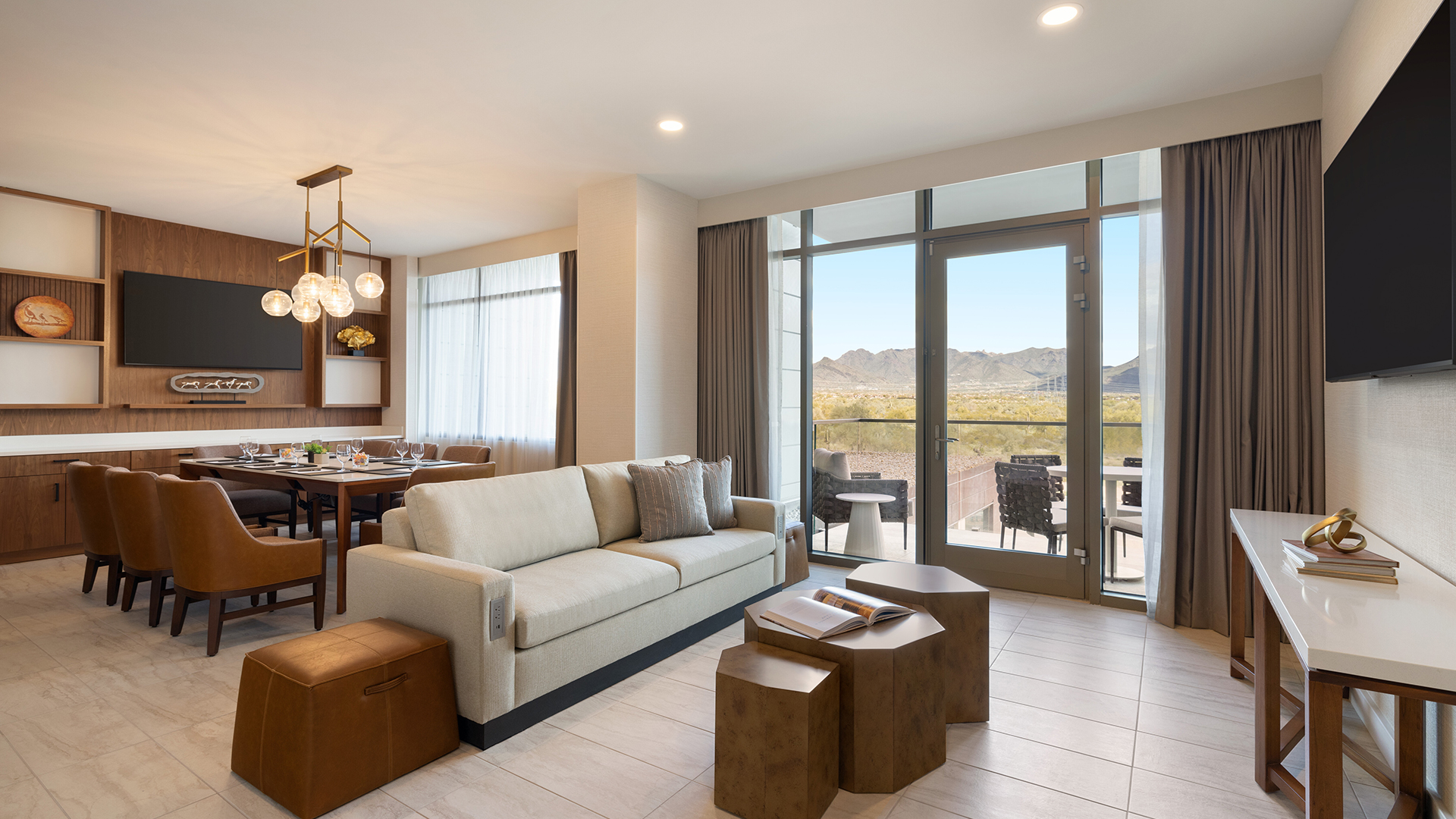 A modern living room features a beige sofa, nested wooden tables, and a wall-mounted TV. The adjacent dining area has a large table with leather chairs. Glass doors open to a balcony with a view of mountains under a partly cloudy sky.