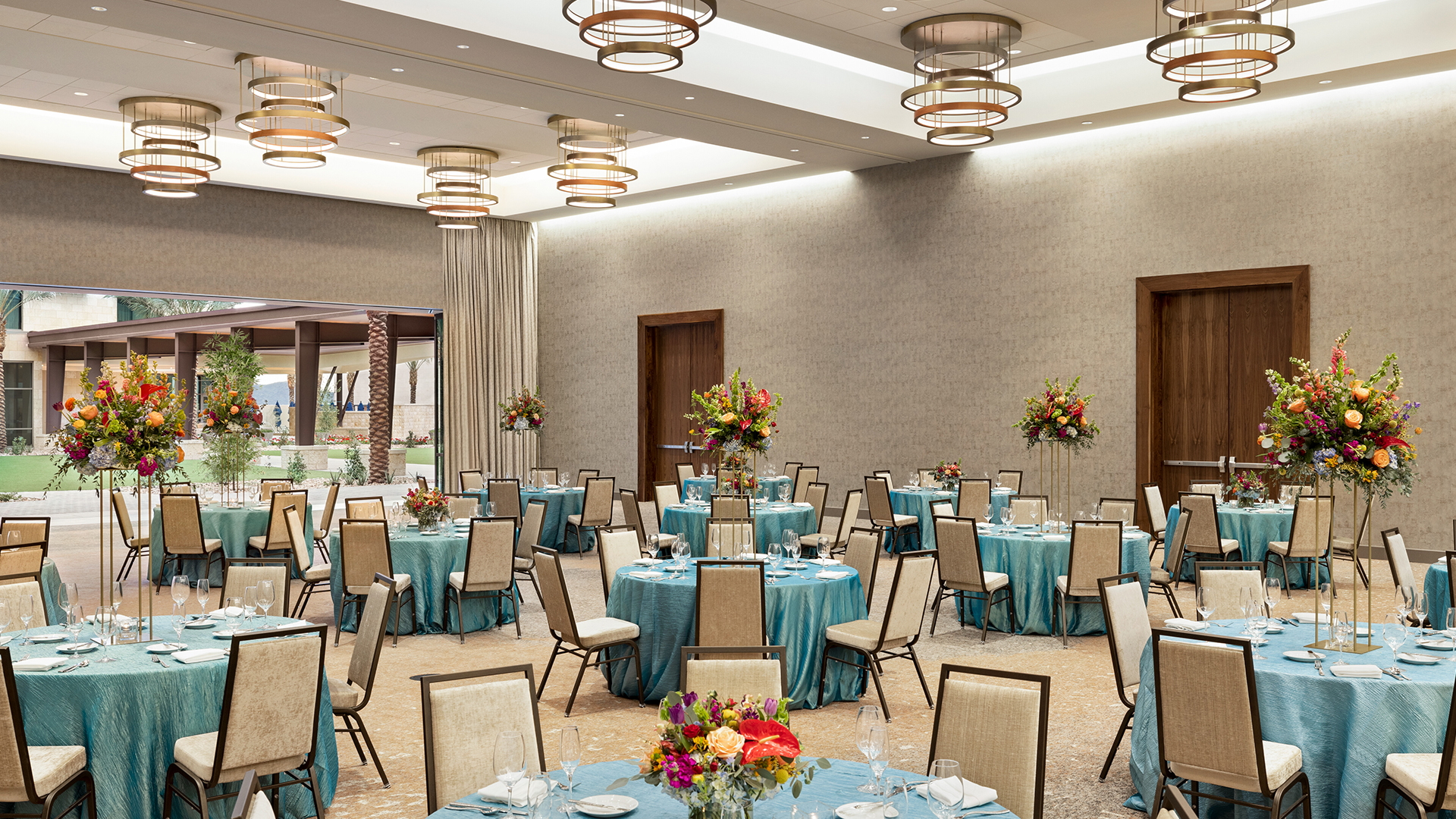 A spacious banquet hall with circular tables covered in turquoise tablecloths, surrounded by beige chairs. Each table has colorful floral centerpieces. Overhead, modern circular light fixtures illuminate the room, and a large window reveals greenery outside.