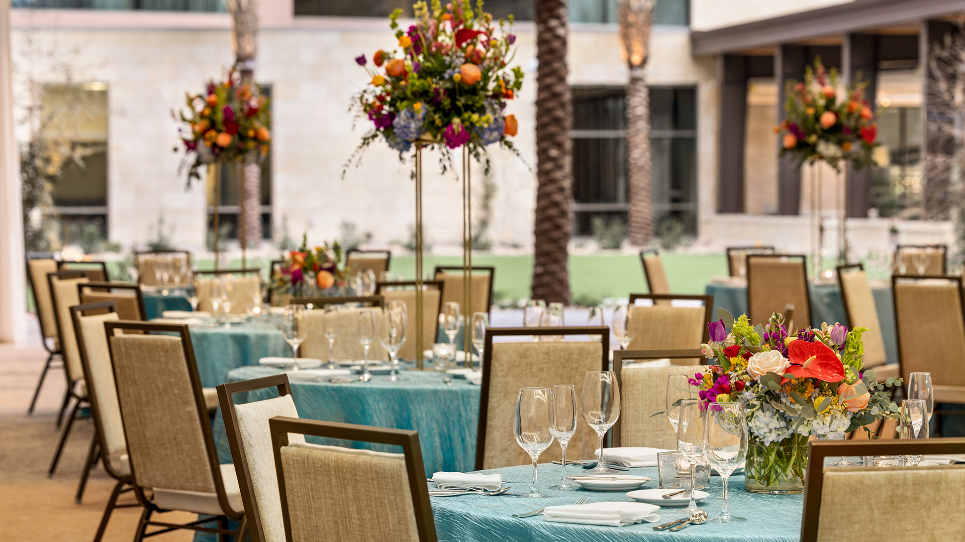 Elegant outdoor dining setup with tables covered in turquoise cloths. Each table is adorned with colorful floral centerpieces and set with glassware and white plates. Tall flower arrangements are in the background under a modern buildings facade.