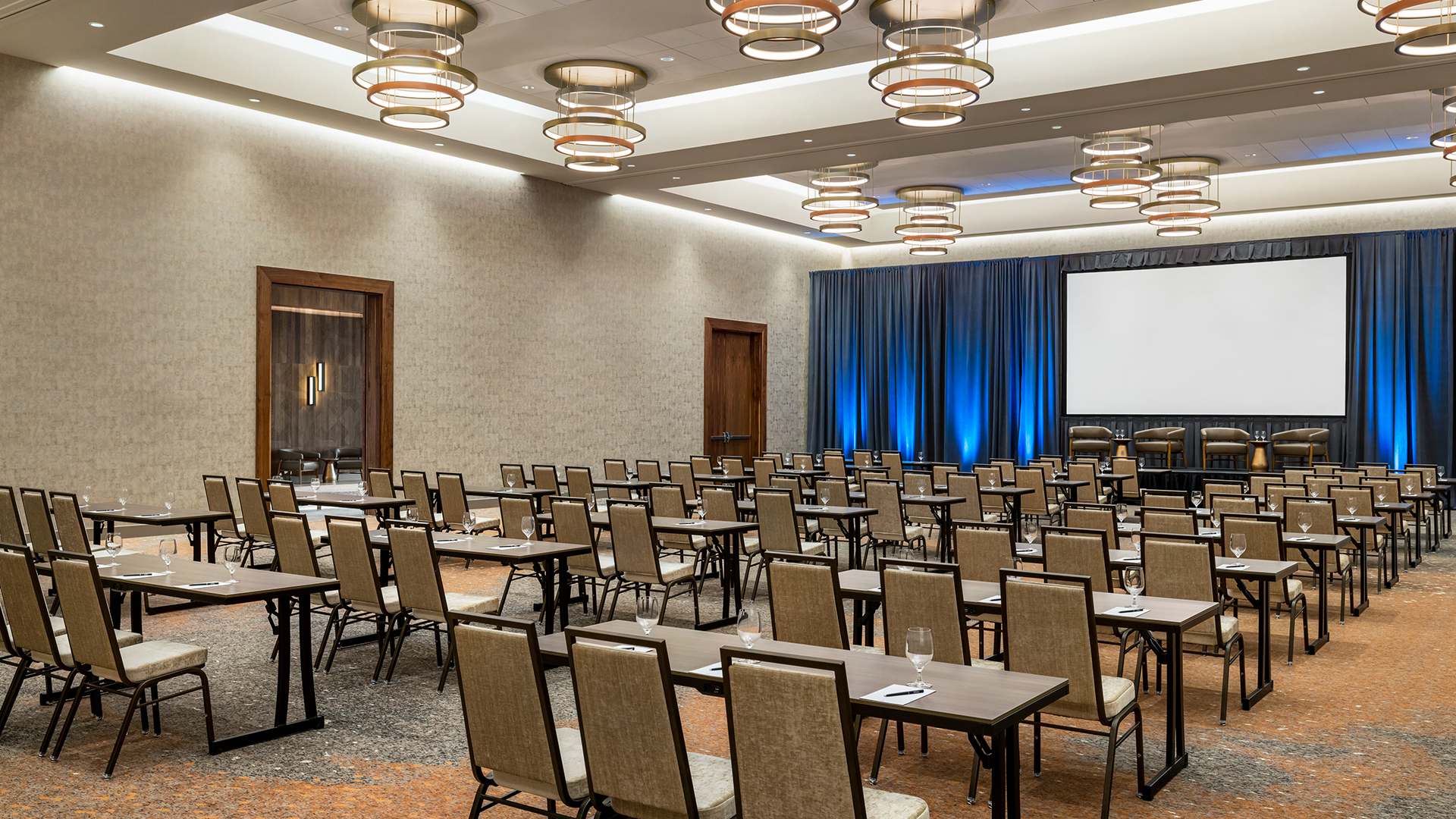 A conference room with rows of chairs and tables facing a large screen on a stage. The room features beige walls, carpeted flooring, and modern circular ceiling lights. Blue curtains hang behind the stage, and glasses are placed on the tables.
