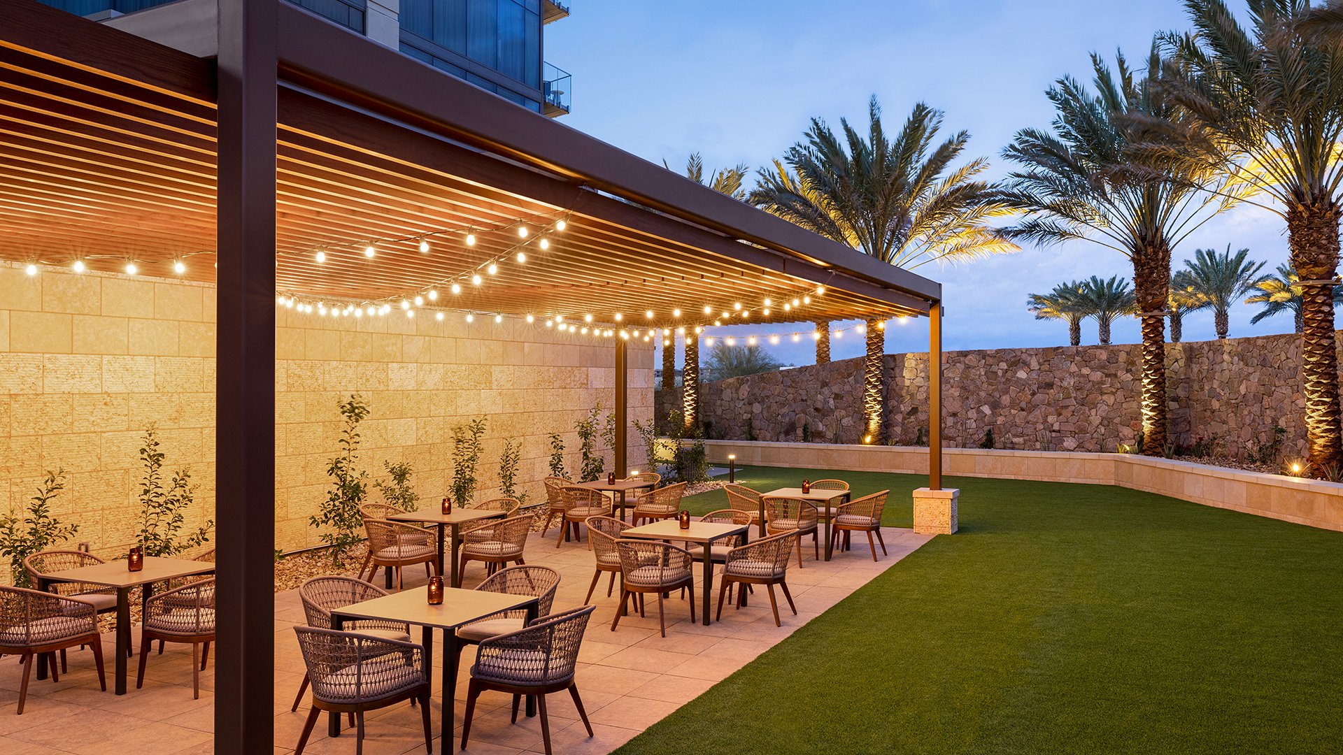 Outdoor dining area at a restaurant with several tables and chairs under a pergola adorned with string lights. Palm trees and a stone wall are in the background, and the area is bordered by well-maintained grass. Twilight sky above.